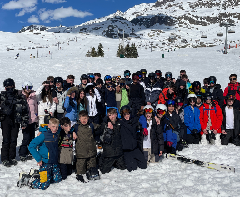 Large group of skiers in helmets and ski gear posing for a group photo on a snowy slope, with chairlifts and mountains in the background.