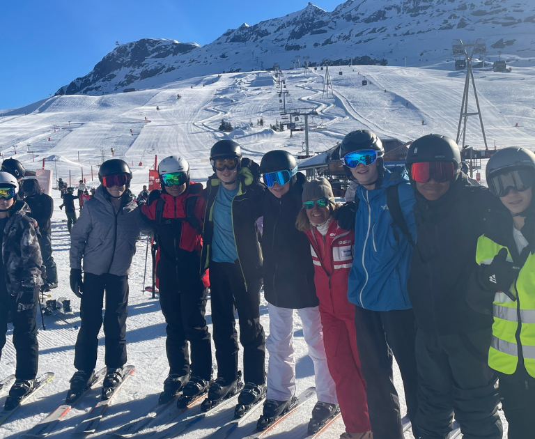 Group of skiers posing in a line on a snowy slope, wearing helmets and colorful jackets, with lifts and mountains behind them on a sunny day.