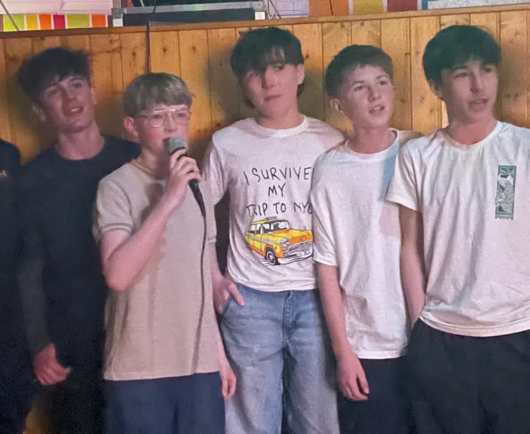 Seven teenage boys stand in a row against a wooden wall, with one holding a microphone and singing while the others look on.