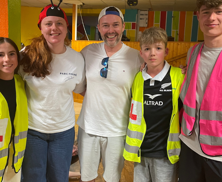 Six people pose for a group photo in a colorful indoor space; several wear neon safety vests while others are in casual clothes.