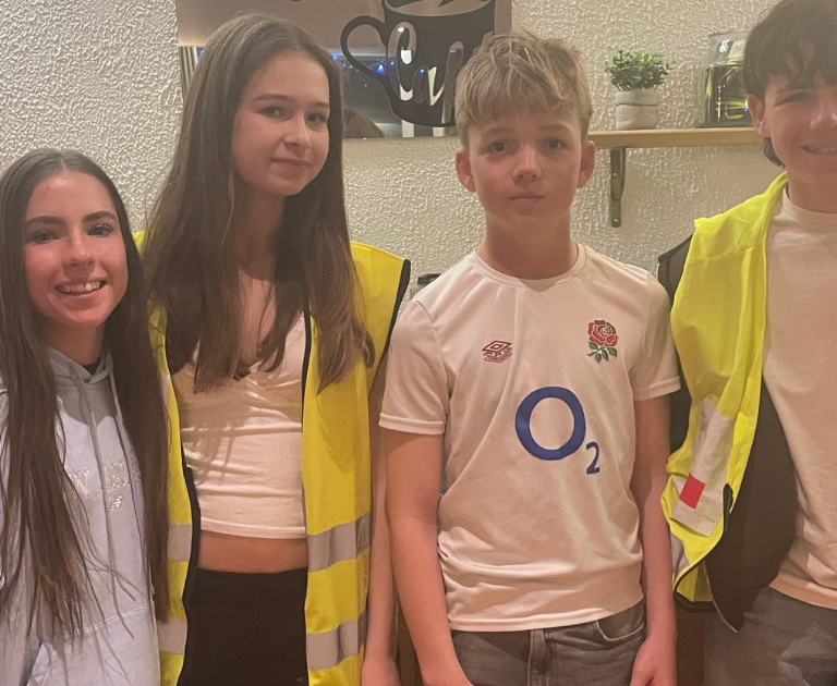 Four teenagers pose and smile indoors, two wearing high-visibility vests, with a boy in a white England rugby shirt in the middle.