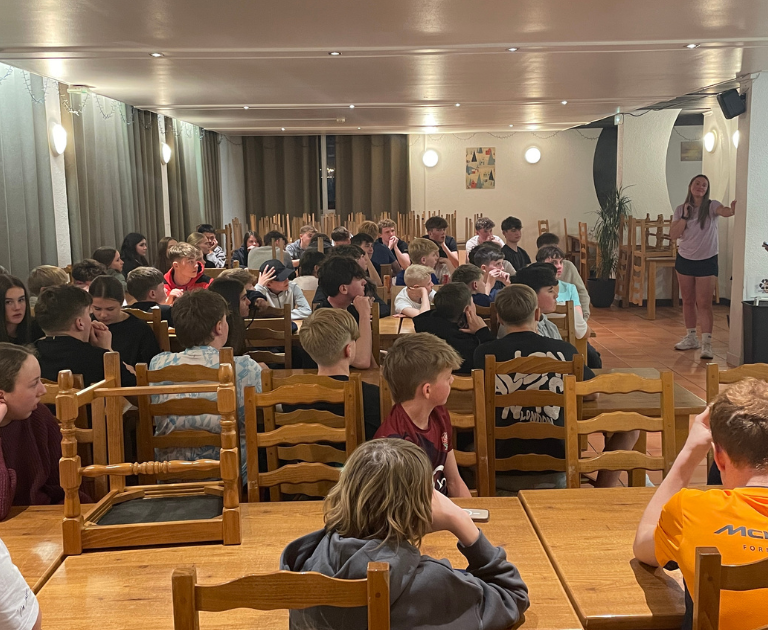 Large group of students seated at long wooden tables in a hall, two presenters stand at the front.