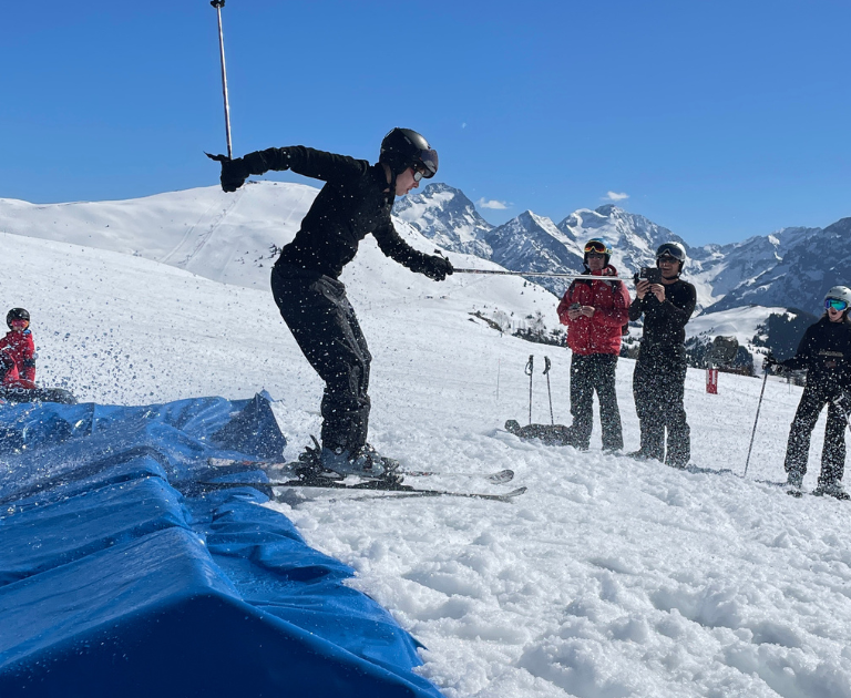 Snowboarder lands on a blue airbag ramp as spectators watch with cameras on a snowy mountain slope under a clear blue sky.