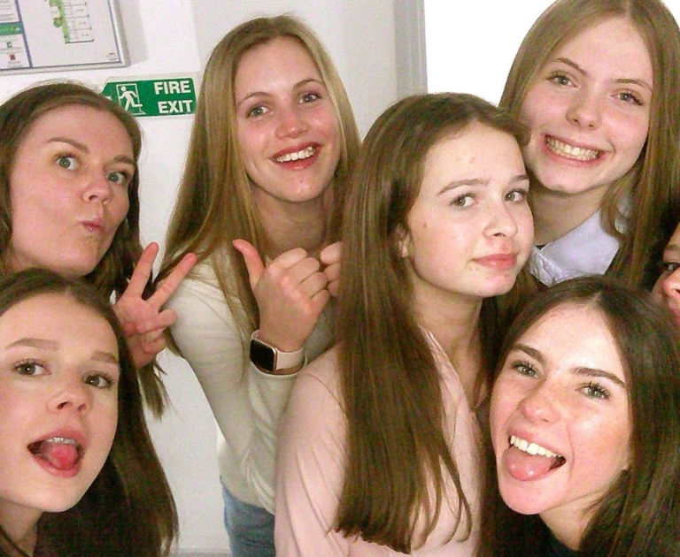 Group of teenage friends in a hallway taking a group selfie, many smiling and sticking out their tongues or posing playfully.