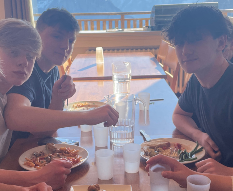 Five teenagers sit around a wooden table on a sunny terrace, eating pasta with vegetables while mountains loom outside.