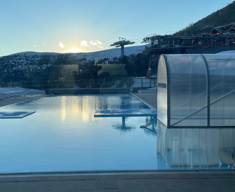 Outdoor pool at a snowy mountain resort during sunset, with a translucent pool enclosure on the right and ski lodges in the background.