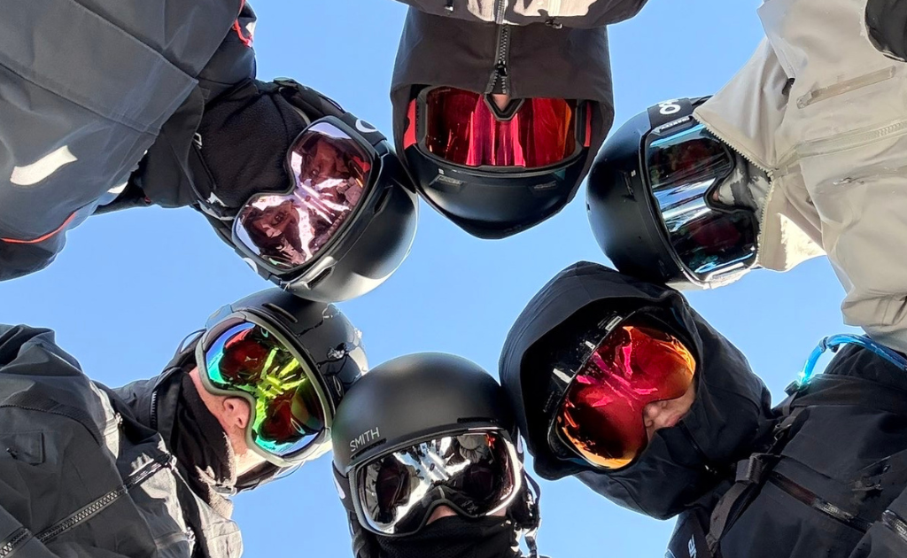 Six skiers/snowboarders wearing helmets and goggles form a circle, looking down at the camera against a blue sky.