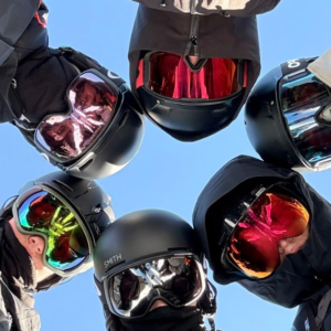 Six skiers/snowboarders wearing helmets and goggles form a circle, looking down at the camera against a blue sky.