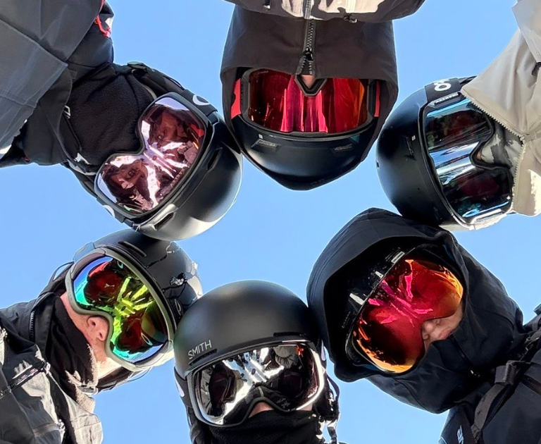 Six skiers/snowboarders wearing helmets and goggles form a circle, looking down at the camera against a blue sky.