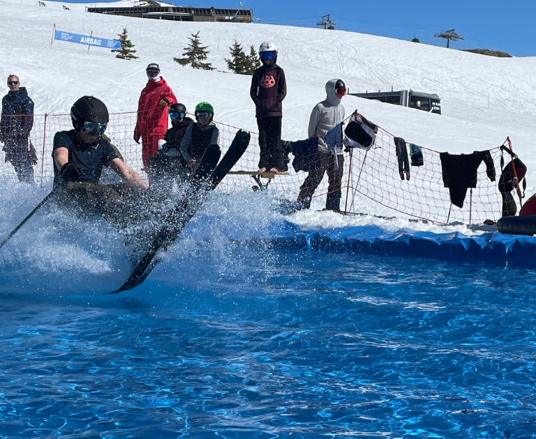 Skier in black gear and helmet crashes into a blue splash pool, sending water spray as onlookers stand behind a safety fence on the snowy slope.