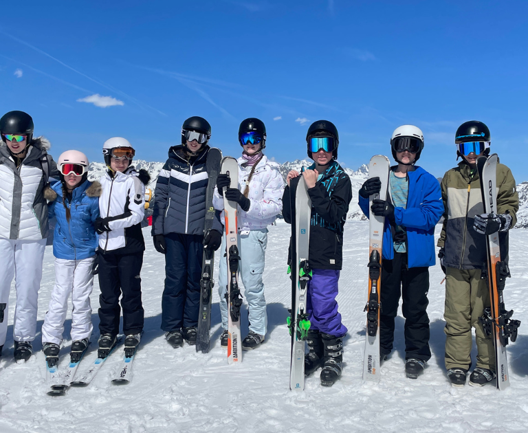Group of eleven skiers in colorful jackets and helmets pose on a snowy mountain with clear blue sky, each holding skis and poles.