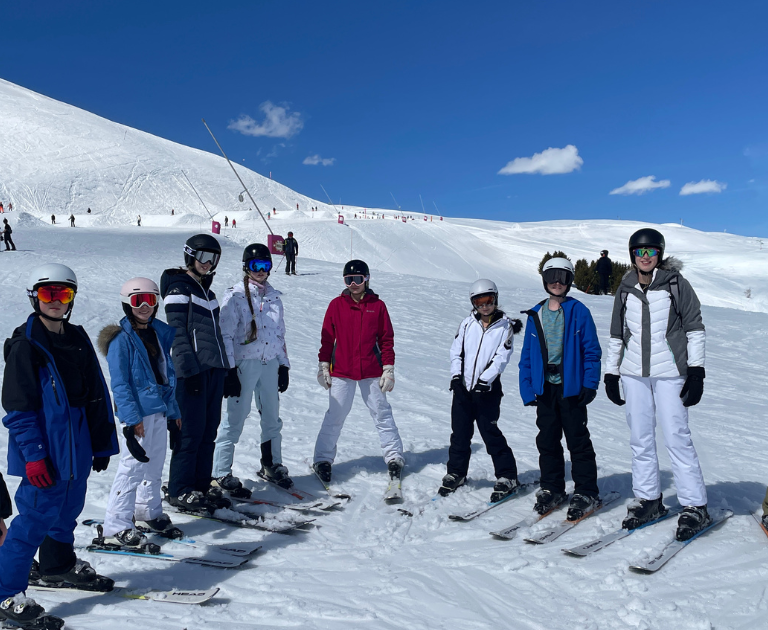 Group of eleven children and teens in ski gear standing in a line on a snowy slope, wearing helmets and goggles under a bright blue sky at a ski resort.
