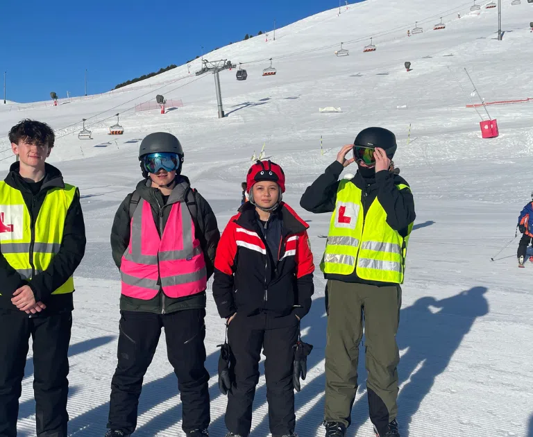 Four skiers wearing bright safety vests pose for a photo on a sunny ski slope with chairlifts in the background.