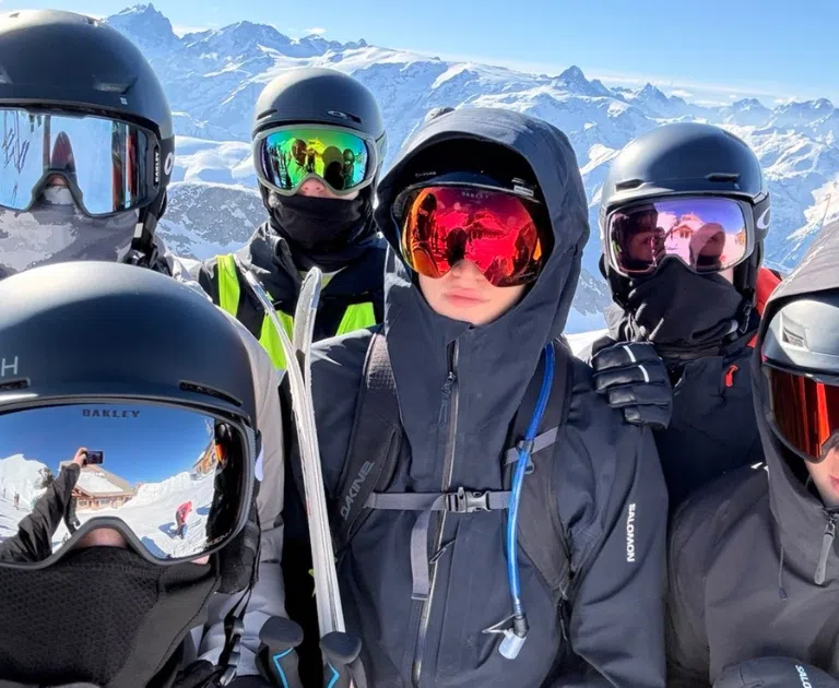 Group of skiers wearing helmets and goggles posing for a selfie with snowy mountains in the background (alpine summit).