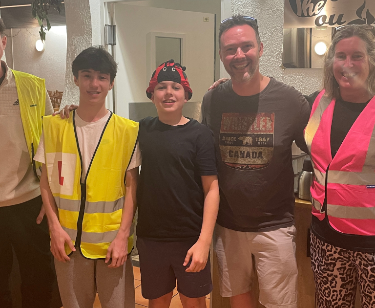 Five people pose together in a kitchen: two teens in yellow safety vests, a younger boy in black, and two adults wearing pink and gray vests.