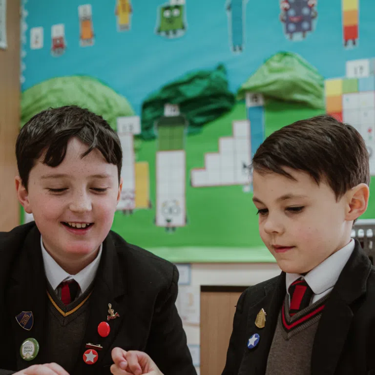Two boys in school uniforms sit at a desk, wearing blazers and ties with badges, smiling as they work in a classroom with bright wall art and charts behind them.