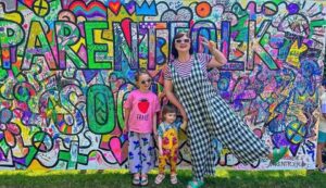 Woman in a black-and-white checkered dress posing with two young girls in front of a bright graffiti wall full of colorful art.