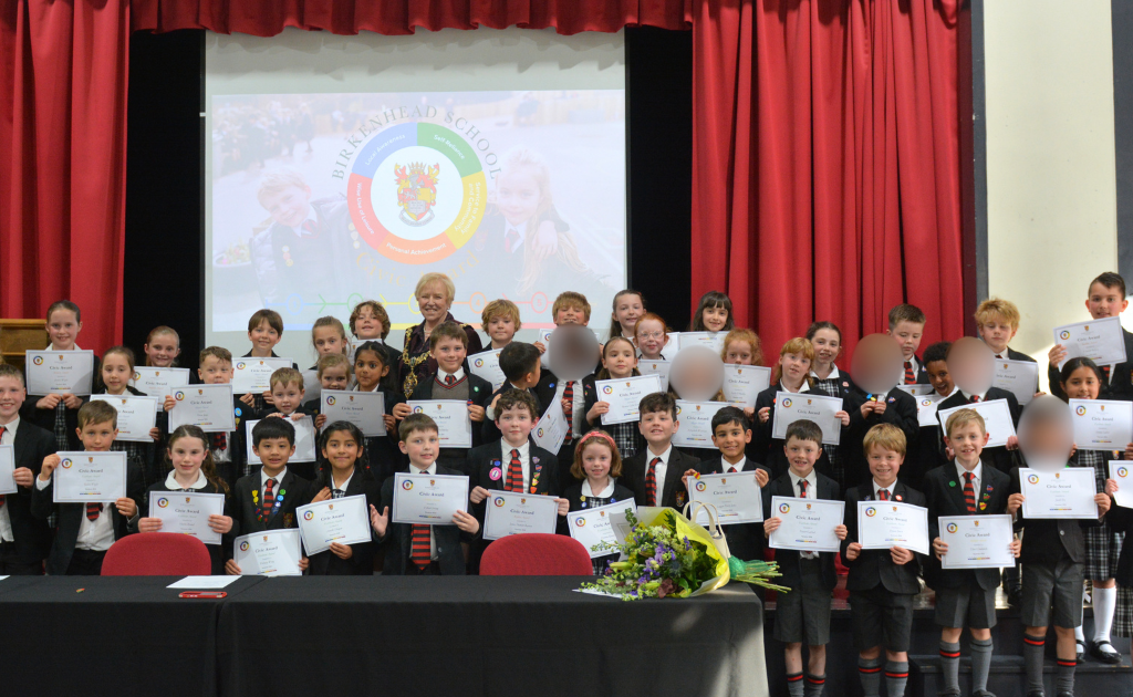 Group of children on stage holding certificates during an awards ceremony; teacher in the center, bouquet on the table, red curtains and a projector screen behind.
