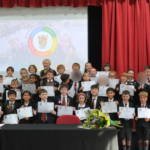 Group of children on stage holding certificates during an awards ceremony; teacher in the center, bouquet on the table, red curtains and a projector screen behind.