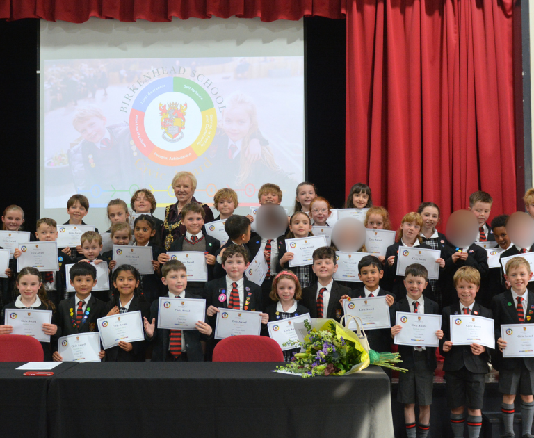 Group of children on stage holding certificates during an awards ceremony; teacher in the center, bouquet on the table, red curtains and a projector screen behind.