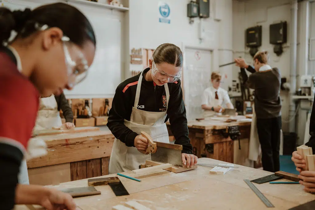 Student in apron and safety goggles planes a piece of wood at a workshop bench in a woodworking class, others working nearby.