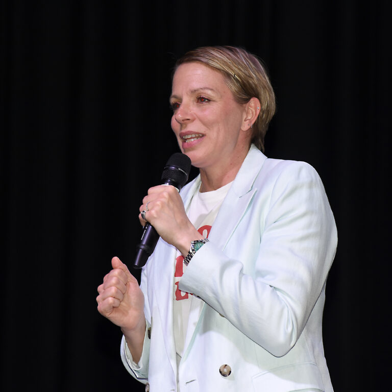 Woman in a light blazer speaks into a handheld microphone on a stage with a black curtain backdrop .