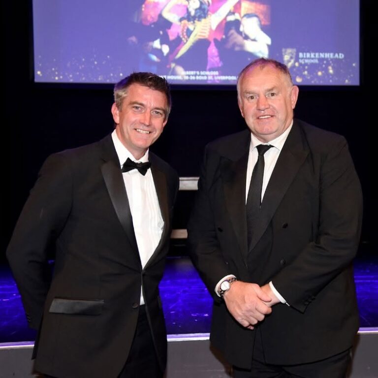 Two men in black tuxedos posing on a stage with a purple-lit floor and a large screen behind them, both smiling at the camera.