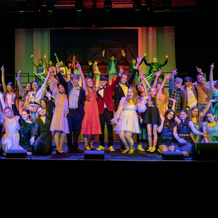 Large group of young performers in colorful costumes posing on a theater stage, raising arms in celebration.