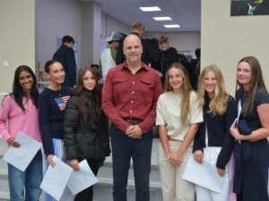 Group of eight people posing for a photo with a man in a red shirt center, students holding papers in a bright hallway event.