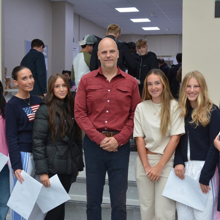 Group of eight people posing for a photo with a man in a red shirt center, students holding papers in a bright hallway event.