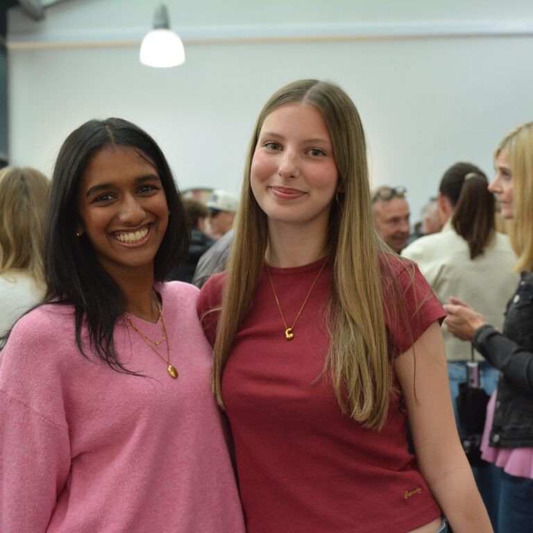 Two young women smile at the camera at a crowded indoor event, wearing pink and maroon tops.