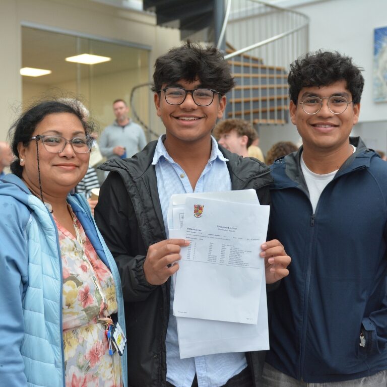 Three people stand together in a school hallway, smiling, with papers in hand.