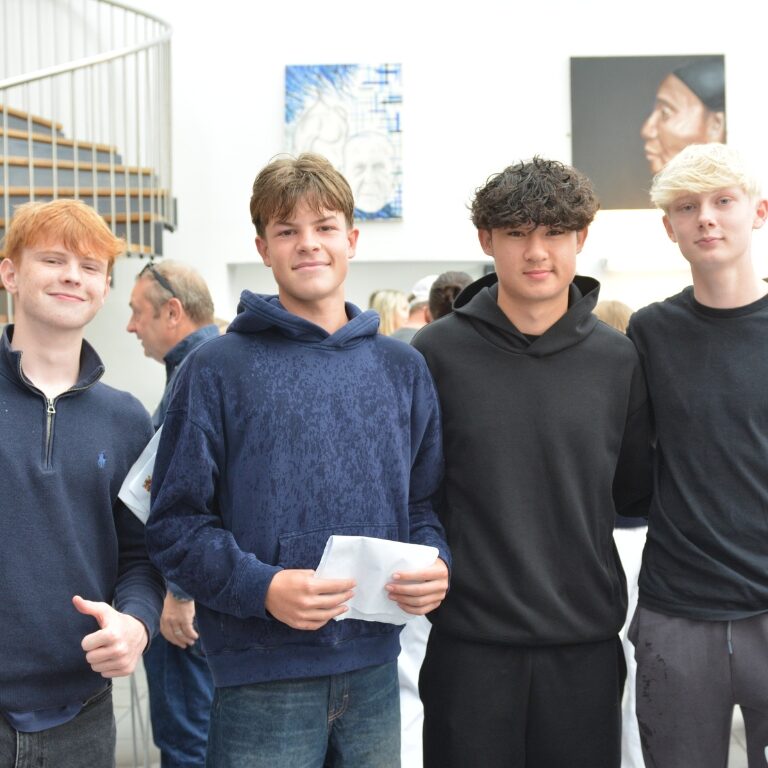 Four teenage boys stand together in a bright gallery, smiling and holding papers nearby.