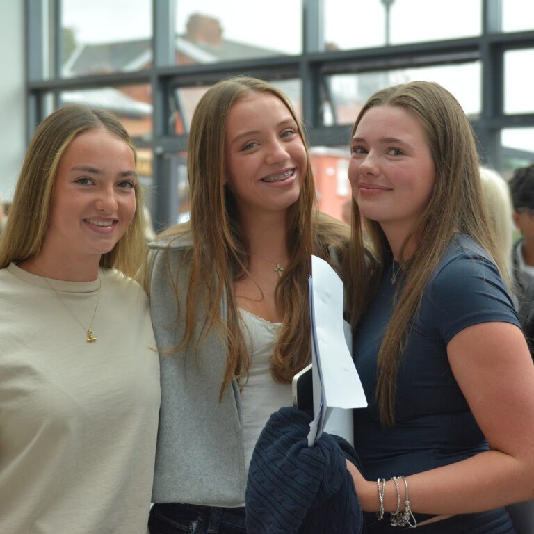 Three smiling teenage girls posing together indoors in a bright, modern space with large windows behind them.