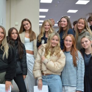 Group of teens posing and smiling for a photo in a well-lit hallway, several holding papers or envelopes as they stand together in casual jackets and sweaters.
