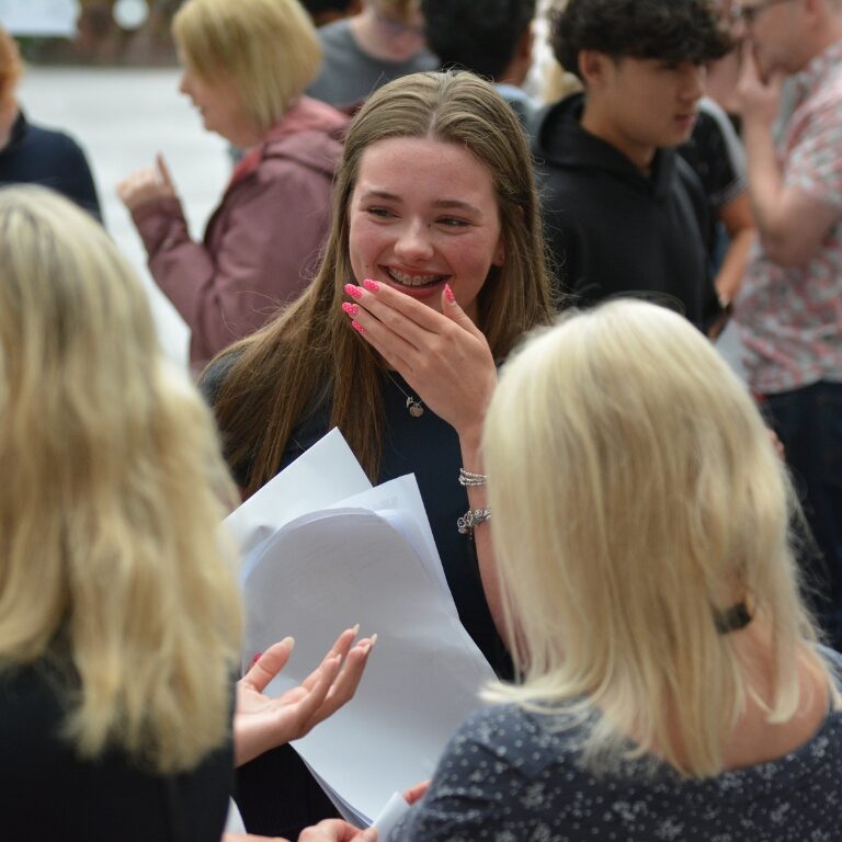 Young woman with long brown hair smiles and covers her mouth while holding papers, amid a crowd.