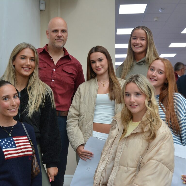 Group of smiling students posing with a bearded man in a red shirt in a hallway.