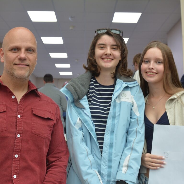Three people pose for a photo in a bright corridor: man in red shirt on the left and two young women smiling at the center and right, one holding a folder.