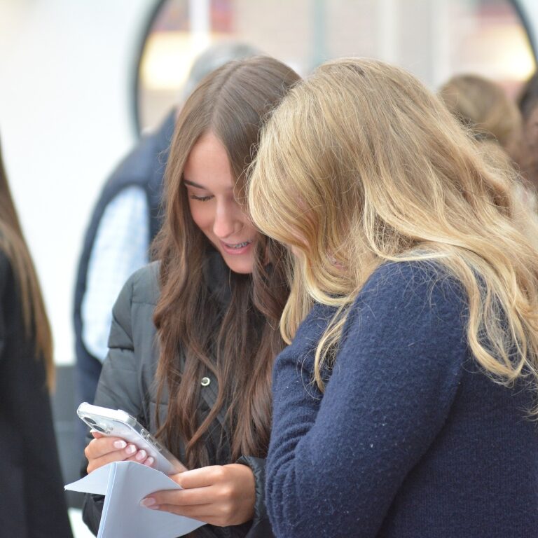 Two young women standing closely, one showing her smartphone to the other while holding papers in a busy indoor space, likely a school or event venue.
