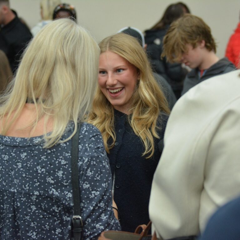 A smiling blonde woman in a dark blue outfit speaks with another person at a crowded social gathering.