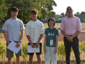 Four people stand outdoors, two young men on left holding papers, a smiling girl with lanyard, and a man in pink shirt on the right, posing for a photo.