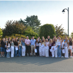 Group of people posing outdoors on a sunny day, standing in two rows with trees and a lamp post in the background.