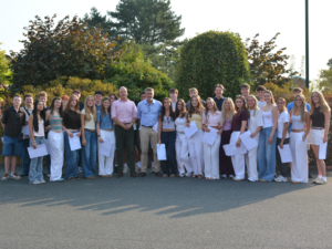 Group of students and two adults posing outdoors for a class photo, many holding papers in front of a hedge.