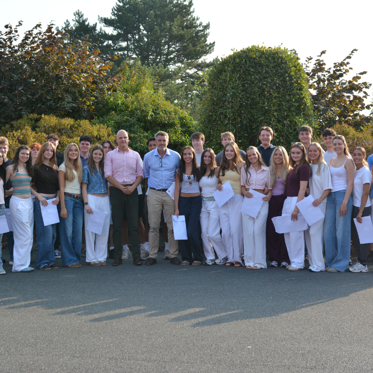 Group of students and two adults posing outdoors for a class photo, many holding papers in front of a hedge.