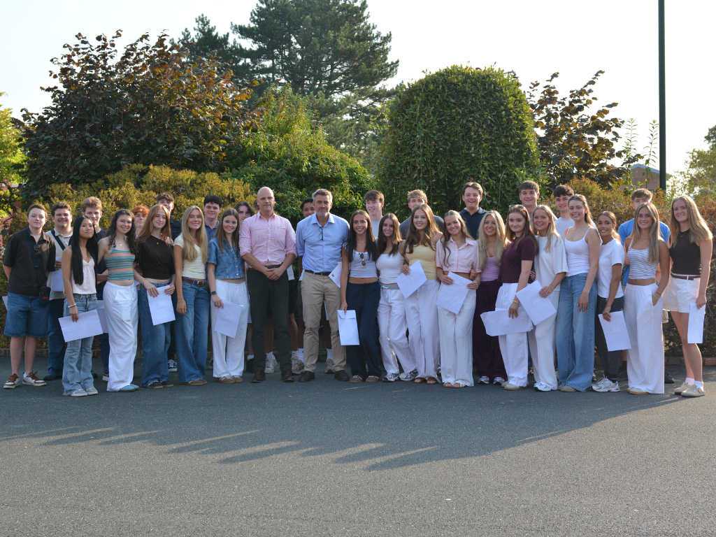 Group of students and two adults posing outdoors for a class photo, many holding papers in front of a hedge.